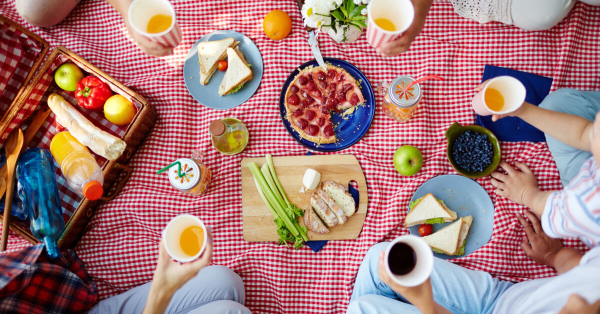 Group of humans with drinks gathered by dinner on picnic cloth